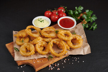 Homemade baked onion rings fries with mayonnaise, Tomato sauce and rosemary on wooden board. fast food products : onion rings on cutting board, on black stone background, unhealthy food.
