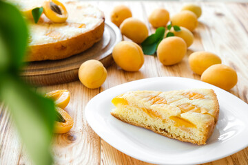 Piece of shortcrust apricot pie on plate with full cake and apricot fruits on background on wooden board. Easy recipe of summer cake for tea time. Homemade bakery by classic recipe.