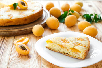Piece of apricot pie on plate with full cake and apricot fruits on background on wooden board. Easy recipe of summer cake for tea time. Healthy nutrition, vegan dessert, gluten-off, sugar free.
