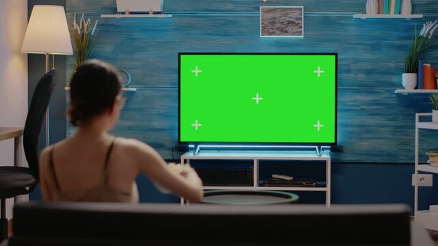Young adult watching green screen on tv at home in modern living room. Caucasian woman having snack on couch, using mockup template display and isolated background for chroma key