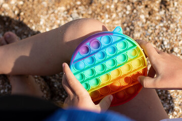 Anonymous child playing with fidget pop it on the beach.New sensory toy for children and adults,top view.Rainbow coloring,summer concept.