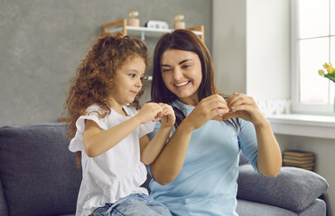 Portrait of little girl and mom sitting on sofa at home, smiling to each other and doing hand heart gesture. Happy woman and daughter sharing positive feelings. Love between mother and child concept