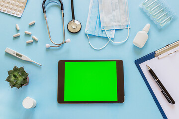 Tablet computer with green screen on blue background. Top view of doctor or nurse desk with various medical supplies and medicines. Flat lay, mock-up.