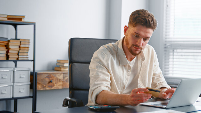 Bearded Businessman In Yellow Shirt Holds Bank Card And Enters Number Information To Laptop