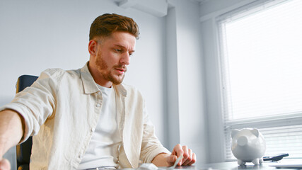 Fototapeta premium Bearded company worker in yellow shirt shakes white piggy bank to listen coins clink over office table