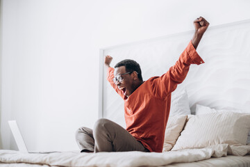 Joyful African-American guy in casual clothes and glasses puts hands up sitting near open laptop on comfortable bed at home