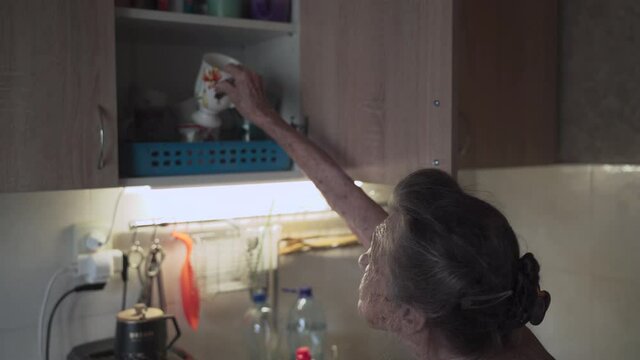 Senior elderly woman washing dishes in kitchen. Sad mature housewife cleans up the old dishes in the kitchen. Upset woman with gray hair 90 years old washing kitchen utensils in a hand wash at home.
