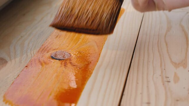 A man paints freshly planed wooden board with a brush on the window background on the workshop. Slow accurate professional wood craftsman work with a gloved hand in the light of a sunny day. Close-up