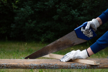 Close up of carpenter wearing gloves and hand sawing wooden plank with old blunt saw in the garden.