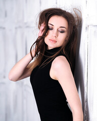 A girl is standing in a small black dress against a background of white panels, the wind blows in her face