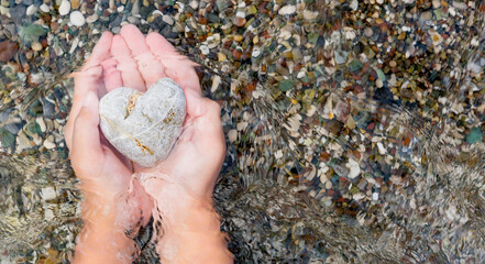 Girl's hands hold a heart-shaped stone in the incoming sea wave. Close-up view. Selective focus. Copy space.