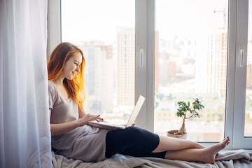 Young attractive brunette sitting on the windowsill, with laptop on her lap, typing on keyboard....