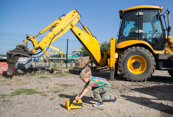 boy child playing with a toy bulldozer on the background of a real big yellow excavator on a sunny summer day. The boy's hobby for construction equipment © Anna