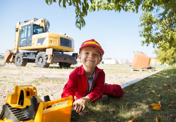 portrait of cute boy, little builder, in a red uniform and cap with toy bulldozer against the...