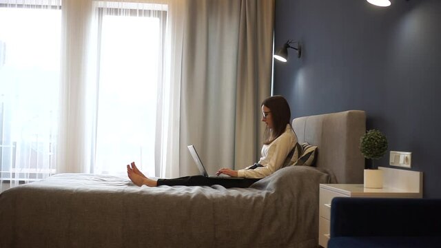 An attractive freelancer girl in glasses with a laptop on the bed in hotel. Travel and work concept with free schedule.