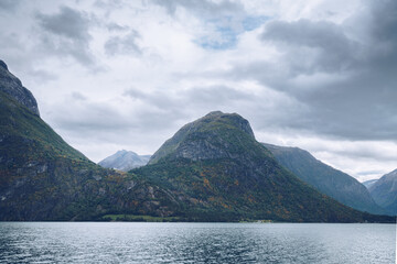 Majestic mountains tower above lake in northern Norway on a cloudy day of Arctic summer. Small houses between the mountains far in the distance. Mysterious Norwegian landscape.