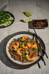 Buckwheat noodles with shrimps, carrots, broccoli, green onions and soy sauce. Vegan soba noodles. Asian food on grey textured table. Seafood healthy diet concept, copy space. 