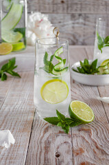 Lemonade with ice, with lime, lemon and mint in a glass and a bottle decorated with cucumber on a skewer on a light background