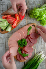 Meat delicacies, cucumbers and tomatoes on a cutting board, on a light background with bread and herbs. A person's hands reach for food
