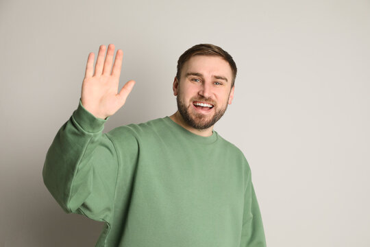 Happy young man waving to say hello on light grey background