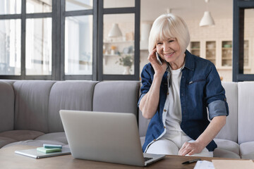 Smiling senior old elderly businesswoman talking on mobile phone while working at home with laptop on couch. Busy old woman having business conversation, communicating with grandchildren. Remote job