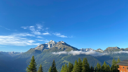 Mountain landscape with snow-capped peaks. Fir trees in the foreground. Clear sky with some white clouds in the background.
