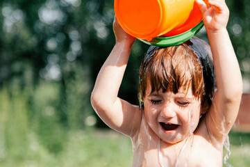 Close-up portrait of a little caucasian laughing girl emotionally pouring water from orange toy bucket on her head in hot summer day. Blurred green background. Happy summertime and childhood concept © makovsvet
