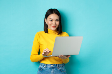 Young asian woman freelancer working on laptop and smiling, standing with computer over blue background - obrazy, fototapety, plakaty