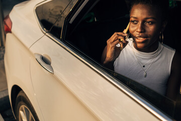 Naklejka premium African American girl talking on smartphone while sitting in the back seat of the car, looking out the window.