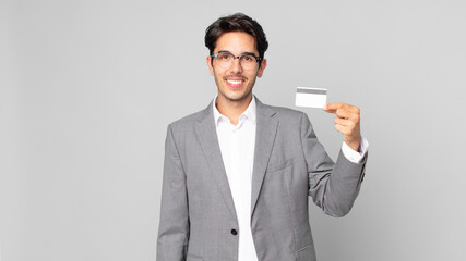 young hispanic man smiling happily with a hand on hip and confident and holding a credit card