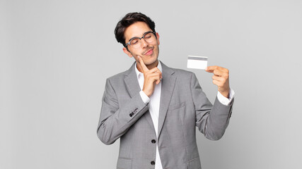 young hispanic man smiling happily and daydreaming or doubting and holding a credit card