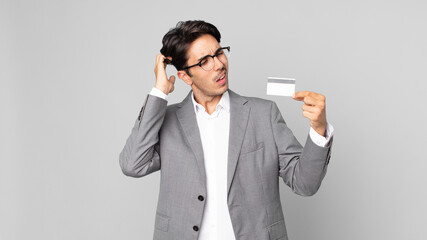 young hispanic man feeling puzzled and confused, scratching head and holding a credit card