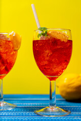 Vertical view of two glasses of the refreshing aperol spritz cocktail with mint and orange leaves on a blue table and yellow background.