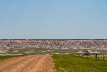 Badlands National Park, SD, USA - June 1, 2008: Landscape with beige geologic deposits appearing from under prairie on horizon under blue sky. Green plains and brown dirt road up front.