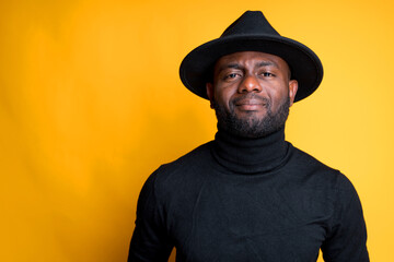 Handsome african american man in hat and black turtleneck looking at the camera while standing on yellow background