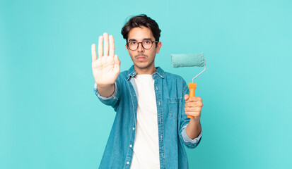 young hispanic man looking serious showing open palm making stop gesture and holding a paint roller