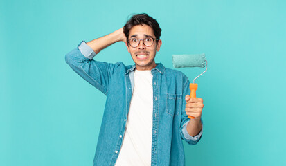 young hispanic man feeling stressed, anxious or scared, with hands on head and holding a paint...