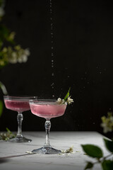 Beautiful glasses on high leg with rose cold cocktail - lemonade and white jasmine flowers, droplets on the white table, dark background cocktail bar concept image with copy space.