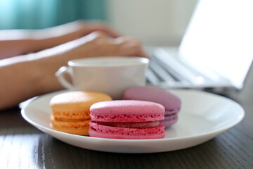 Macaroons in white plate on background of woman working at laptop. Almond cookies with coffee cup, eating sweets at work