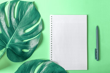 Fototapeta na wymiar Monstera leaves, checkered notebook and pen on light green background. Copy space. Close-up