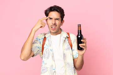 young hispanic man feeling confused and puzzled, showing you are insane and holding a bottle of beer