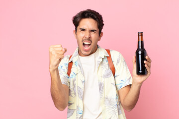 young hispanic man shouting aggressively with an angry expression and holding a bottle of beer
