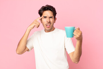 young hispanic man feeling confused and puzzled, showing you are insane and holding a coffee mug