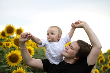 brunette woman young mom with cute baby blonde son on a field of sunflowers.