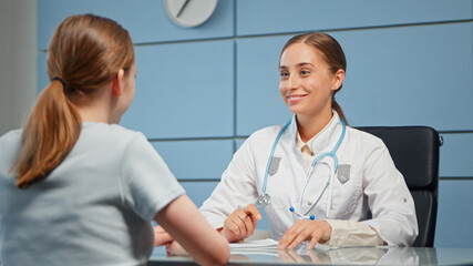 Smiling blonde general practitioner in white coat with blue stethoscope consults young woman with long hair