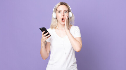young pretty albino woman feeling shocked and scared with headphones and smartphone