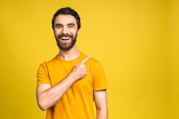 Look over there! Happy young bearded handsome man in casual pointing away up and smiling while standing isolated over yellow background.
