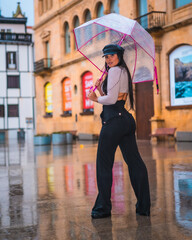 Posing of a young brunette Latina with a leather cap in the autumn rain with a transparent umbrella in the city