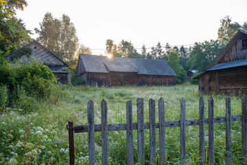 Old, damaged, wooden agricultural buildings situated between green trees, on a meadow. Forest in the background, wooden fence in the foreground. Krasnobród, Roztocze, Poland.