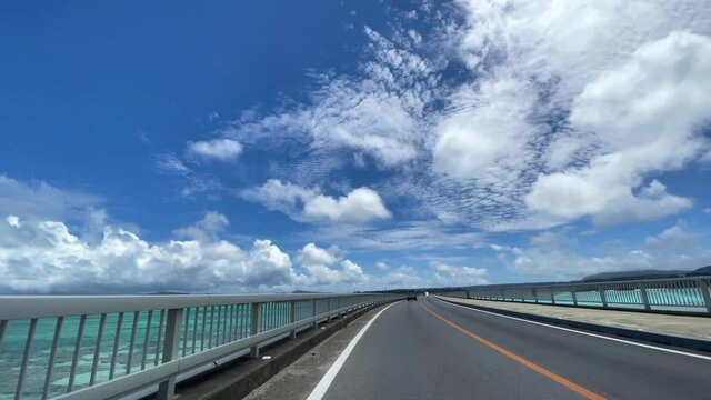 OKINAWA, JAPAN - JUNE 2021 : Driving at Kouri Island bridge. Wide camera point of view (POV), seaside road driving shot. Blue sunny summer sky.